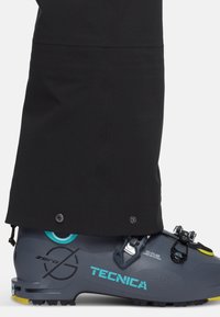 Γκρίζα μπότες σκι με τυρκουάζ και κίτρινα διακοσμητικά, λεπτομερείς πόρπες και υφή στην επιφάνεια, μερικώς καλυμμένες από το παντελόνι σε μαύρο χρώμα.