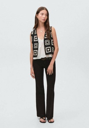 Schwarze und weiße gehäkelte Weste mit quadratischen Mustern über einem weißen Oberteil, kombiniert mit weit ausgestellten schwarzen Jeans und schwarzen Sandalen.