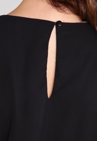 Vêtement noir avec une ouverture en forme de goutte d'eau à l'arrière, agrémentée d'un petit bouton noir au niveau du col. Texture du tissu lisse.