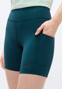 Κοντινό πλάνο ενός ατόμου που φοράει αθλητικά σορτς σε τυρκουάζ απόχρωση με πλαϊνή τσέπη όπου κρατά το χέρι του, με ένα μικρό μαύρο λογότυπο στον μηρό.