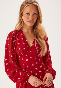 Abito floreale rosso realizzato in tessuto leggero con scollatura a V profonda e maniche lunghe a sbuffo, caratterizzato da motivi floreali bianchi e dettaglio in vita arricciato.