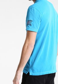 Polo shirt bleu clair à manches courtes avec une coupe décontractée. Présente un logo brodé sur la manche gauche et un ourlet droit avec des fentes sur les côtés.