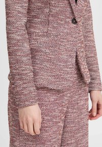 Strukturierter Blazer in rot-weißer Webart; verfügt über einen einzelnen schwarzen Knopf und seitliche Taschen. Lange Ärmel mit taillierten Manschetten.