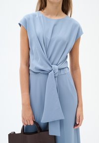 Abito azzurro chiaro con maniche corte, caratterizzato da un dettaglio a nodo torcigliato in vita. La modella tiene una borsa a tracolla di color marrone scuro. Tessuto dalla texture liscia.