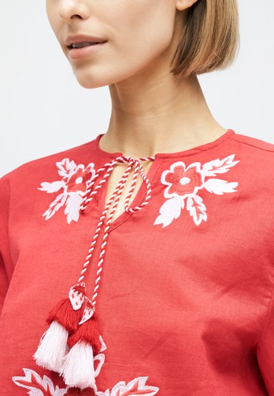 Femme portant un chemisier rouge avec une broderie florale blanche et des cordons torsadés se terminant par des pompons rouges et blancs au niveau de l'encolure.