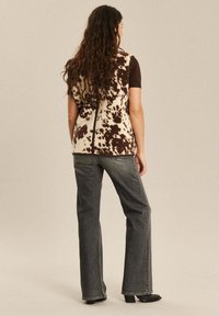 Weste mit braunem und cremefarbenem Muster aus strukturiertem Stoff, kombiniert mit dunkelgrauen Schlagjeans und schwarzen Stiefeletten mit Absatz.