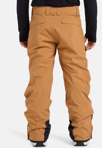 Braune wasserdichte Hosen im lässigen Schnitt, mit seitlichen Reißverschlüssen an den Knöcheln und einem strukturierten Stoff. Zwei Rückentaschen sind sichtbar.