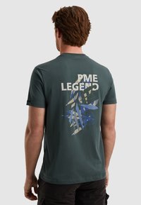 Grøn bomulds T-shirt med korte ærmer, der har et grafisk design på bagsiden, som inkluderer "PME LEGEND" og blå abstrakte elementer.