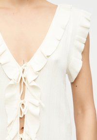 Γυναίκα που φοράει λευκό αμάνικο τοπ με βολάν στις άκρες και κουμπώματα με δέσιμο στο μπροστινό μέρος, με ορατό μέρος του λαιμού και του άνω μέρους του κορμού της.