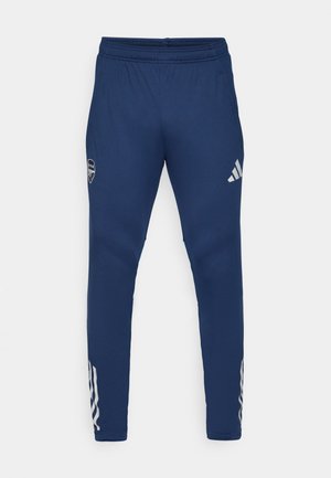 Granatowe spodnie sportowe z elastycznym pasem, zwężanym krojem nogawki, srebrnym logo na udzie oraz charakterystycznymi trzema paskami na dolnej części nogawki.