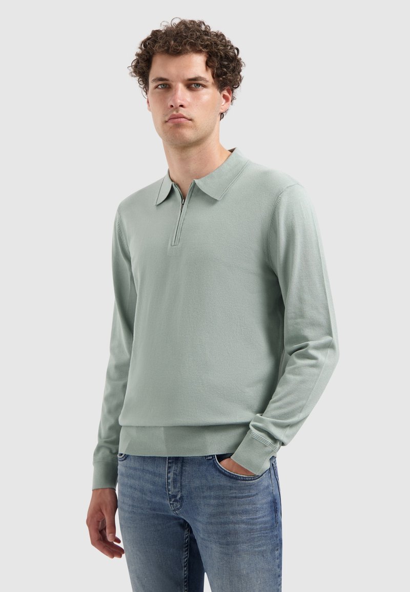Lichtgroene long sleeve polo met een ritssluiting bij de kraag, textuurstof, geribbelde manchetten en een ontspannen pasvorm. Gecombineerd met blauwe jeans.
