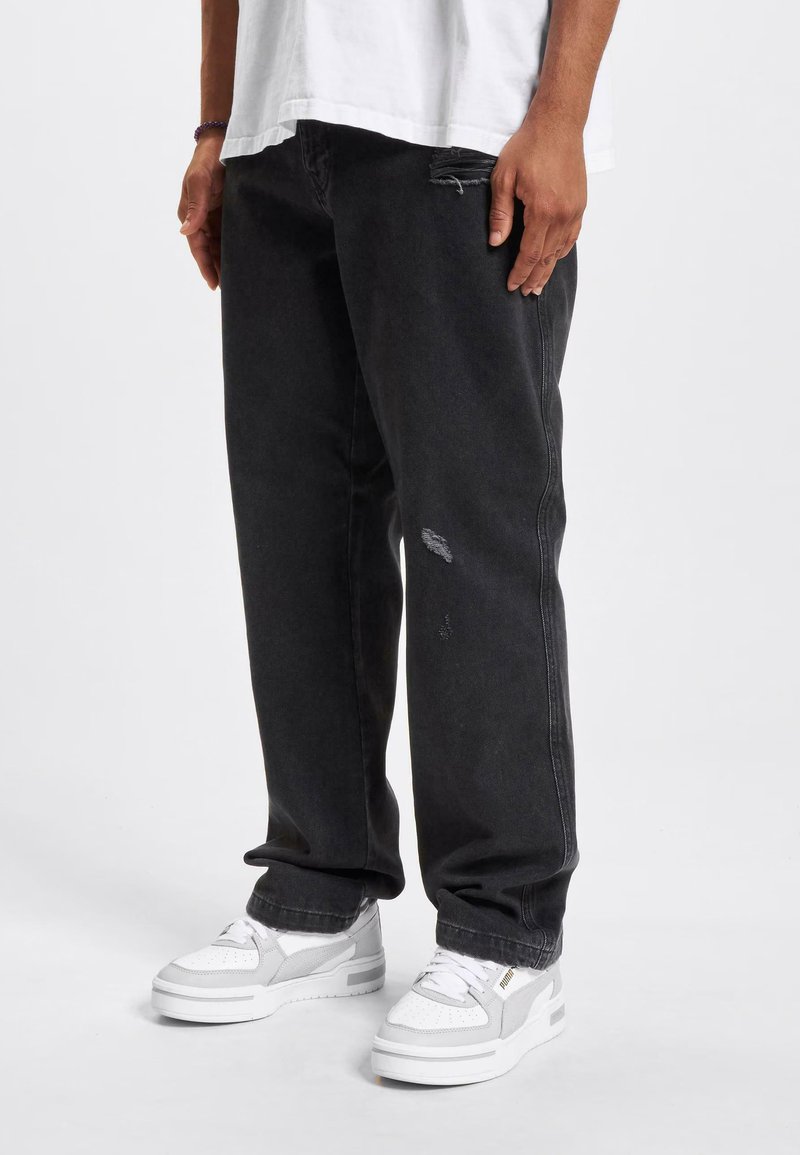 Jeans de mezclilla negra con un corte relajado, que presentan detalles desgastados y costura visible. Combinados con zapatillas blancas y una camiseta blanca holgada.