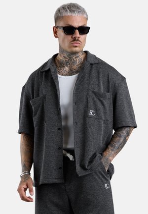Mann mit hellem Haar und Hals-Tattoos, trägt schwarze Sonnenbrille, ein gemustertes graues Hemd- und Hosenset mit weißem Tanktop darunter.
