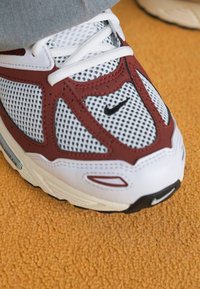 Nike sporta apavi ar baltu sietu augšdaļu un bordo akcentiem, ar noapaļotu purngalu, biezu zoli un teksturētām detaļām.