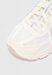 Sapatilha atlética branca com materiais em malha e couro, apresentando um design texturizado, atacadores e sola de borracha. Os detalhes incluem sutis toques de cinza.