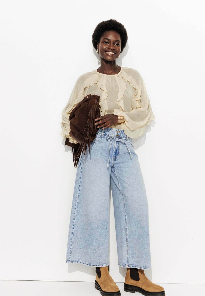 Blouse beige clair avec des bords volants, culottes en denim bleu surdimensionnées, sac en suède brun avec des franges, et bottines beige avec des semelles noires.