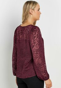 Blusa trasparente bordeaux con maniche lunghe, stampa animale testurizzata, scollatura rotonda e dettagli arricciati sulle spalle.