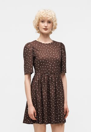 Frau mit kurzen lockigen blonden Haaren trägt ein braunes Kleid mit weißen Punkten, aufgepufften kurzen Ärmeln und eingesetzter Taille, steht vor weißem Hintergrund.