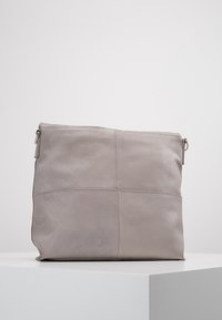 Šedá kožená taška se čtvercovým designem, s hladkým povrchem a jemnými švy. Zapínání na zip, stojí vzpřímeně na povrchu.