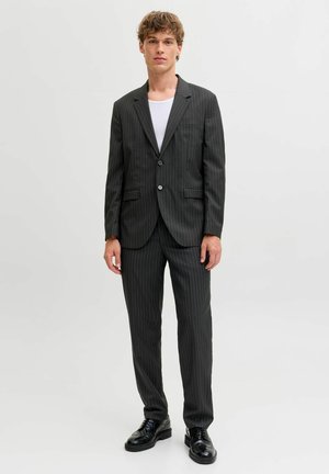 Egy fiatal férfi áll, sötét csíkos öltönyt viselve, fehér inggel és fekete elegáns cipővel, egyszínű világos háttér előtt.