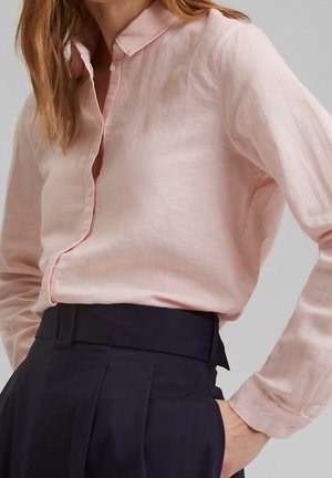 Kobieta w jasnoróżowej, długorękawowej bluzce zapinanej na guziki, włożonej w granatowe, plisowane spodnie z wysokim stanem, z ręką w kieszeni.