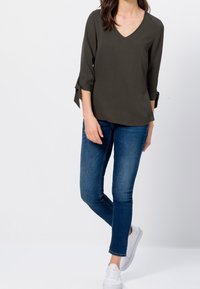 Dunkelgrünes V-Ausschnitt-Blusenshirt mit aufrollbaren Ärmeln, kombiniert mit enganliegenden blauen Jeans und weißen Turnschuhen. Glatte Textur und lässiges Design.