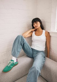 Gröna mockasneakers med lila detaljer, ljusblå avslappnade jeans, och en vit ribbad linne. Sitter på en grå klädd soffa.