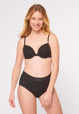 Kvinde smilende, iført sort bh og sorte trusser med høj talje, stående mod en ensartet lys baggrund.