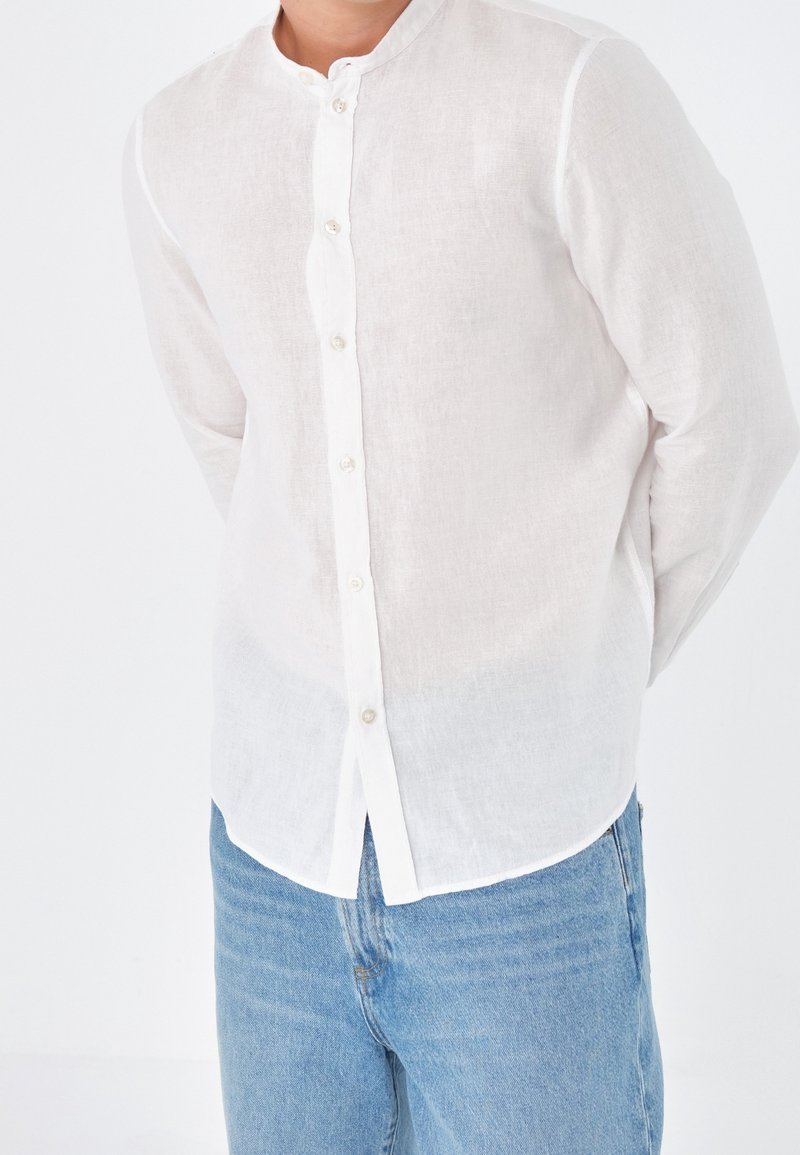 Chemise blanche en lin à boutons avec un col mao, dotée d'une patte de boutonnage sur le devant et d'un ourlet arrondi, associée à un jean en denim bleu clair.