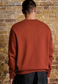 Homme aux cheveux courts et portant une boucle d'oreille, vêtu d'un sweat-shirt couleur rouille et d'un jean foncé, se tenant face à un mur en briques texturé.