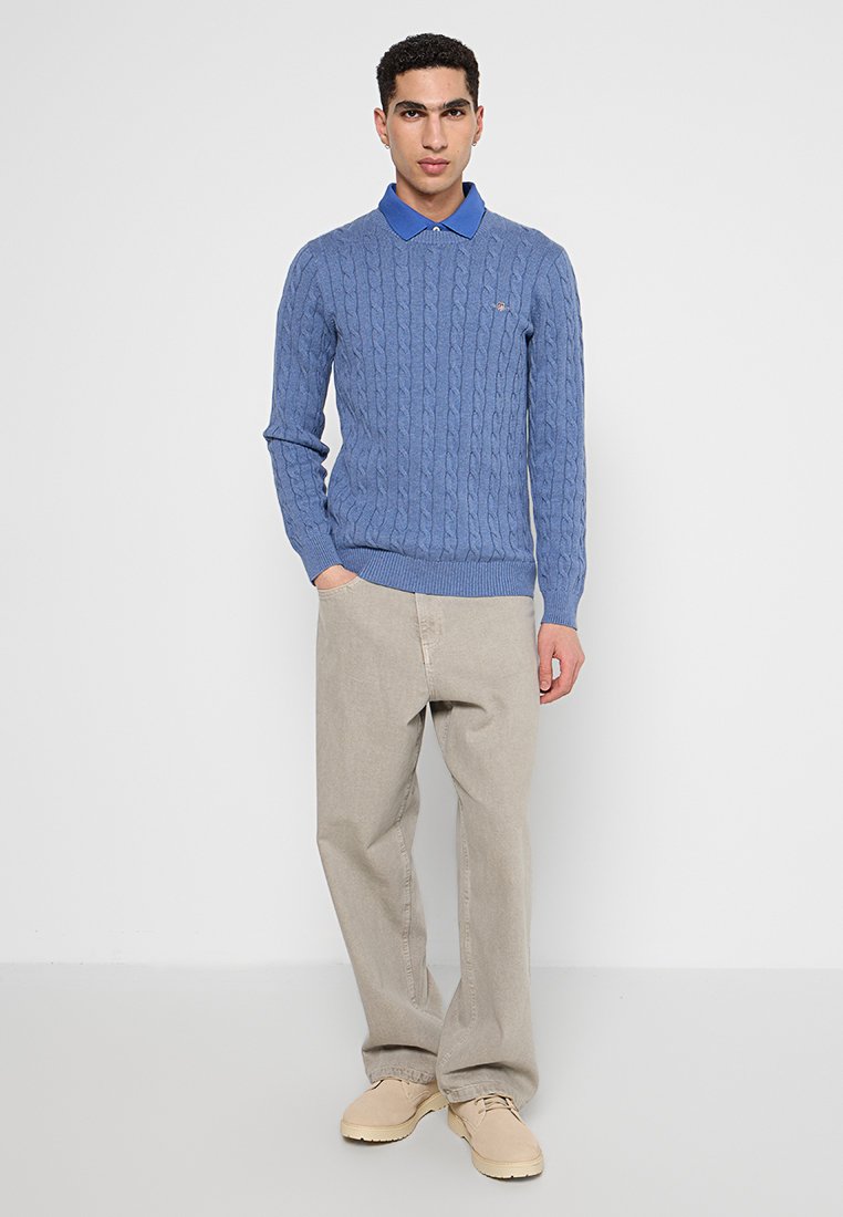 Suéter de punto azul con un patrón de trenzado, puños y dobladillo acanalados, combinado con pantalones beige y zapatos claros, con una camisa azul con cuello debajo.