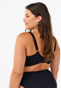 Zwarte bh met verstelbare banden, voorzien van metalen accenten en een smooth textuur. Model van achteren afgebeeld, in combinatie met bijpassende slip.