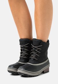 Botas de invierno negras hasta el tobillo con suelas grises, con cierre de cordones y un ligero tacón, llevadas sobre piernas desnudas frente a un fondo blanco.