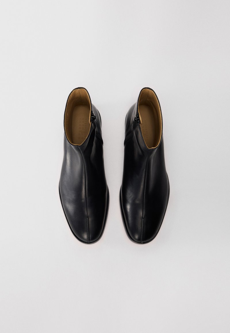 Une paire de bottines en cuir noir avec des zips sur les côtés, doublure intérieure beige, présentée de dessus sur un fond blanc uni.