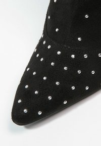 Schwarzer Wildleder-Stiefelette mit spitz zulaufender Spitze, verziert mit gleichmäßig verteilten silbernen Nieten auf dem Obermaterial für eine strukturierte Optik.