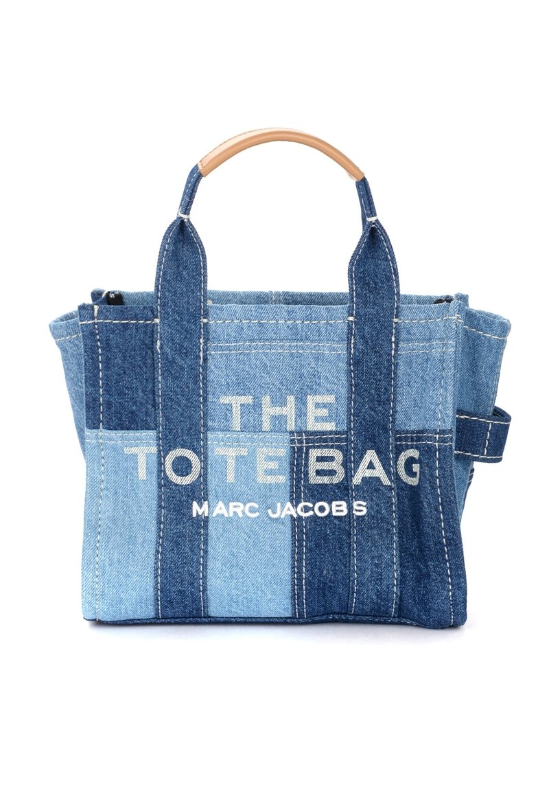 Denim tote bag with a blue color gradient, featuring white text that reads "THE TOTE BAG" and "MARC JACOBS," and wood-like handles.