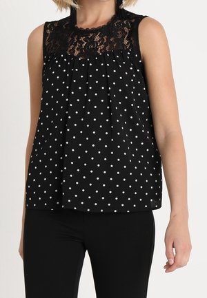 Czarna bluzka bez rękawów w białe kropki z koronkową wstawką na ramionach, noszona z czarnymi spodniami, widoczna górna część tułowia i ramię.