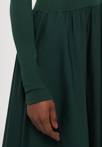 Dark green dress with long sleeves, a fitted bodice, and a flowing, gathered skirt. Smooth texture and simple design.