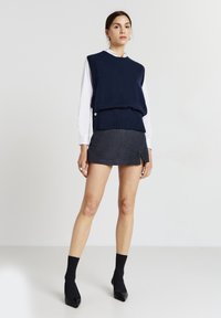 Pull sans manches en tricot marine superposé sur une chemise blanche, associé à une mini jupe gris foncé, des chaussettes noires et des chaussures noires à bout pointu.