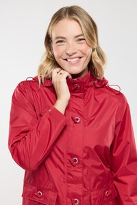 Femme souriante aux cheveux blonds portant un imperméable rouge, tenant le col de sa main gauche contre un fond uni.