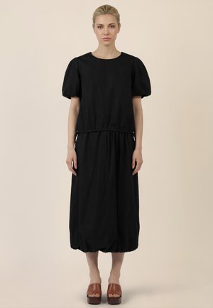 Frau mit blonden Haaren, die ein schwarzes Kleid mit Puffärmeln und braune Plateausandalen trägt, steht vor einem beigen Hintergrund.