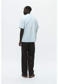 Chemise bleu clair à manches courtes avec une coupe droite, associée à un pantalon sombre et ample, et des chaussures marron, vue de dos.
