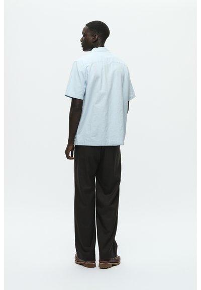 Chemise bleu clair à manches courtes avec une coupe droite, associée à un pantalon sombre et ample, et des chaussures marron, vue de dos.