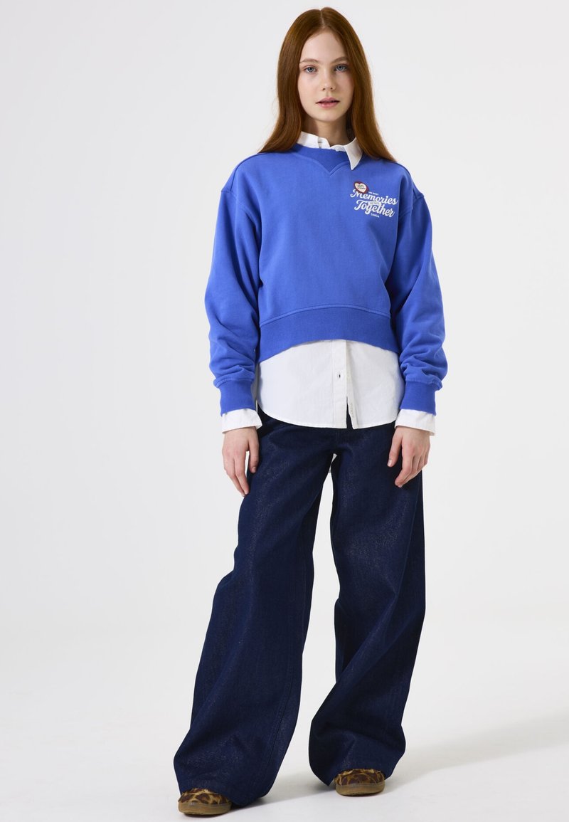 Sudadera azul corta con texto bordado; superpuesta sobre una camisa blanca de botones. Jeans oscuros de corte holgado y calzado con estampado.