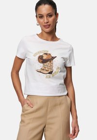 Hvid bomulds T-shirt med et grafisk design af cowboy støvler og en hat med teksten "SHERIFF HJEMME" i gul, omgivet af stjerne accenter.