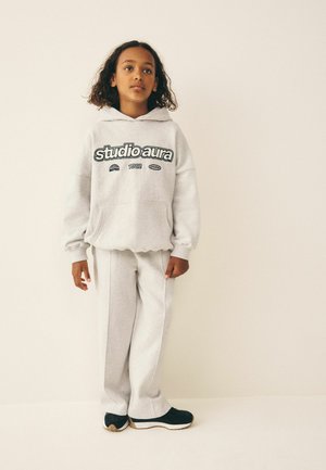 Young person standing against a plain background wearing a light gray "studio aura" hoodie, matching pants, and black sneakers.