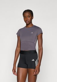 Lühikeste varrukate ja ümmarguse kaelusega lilla sportlik crop top halli logoga, koos mustade lühikeste pükste ja valgete triipudega, millel on veniv vöökoht.