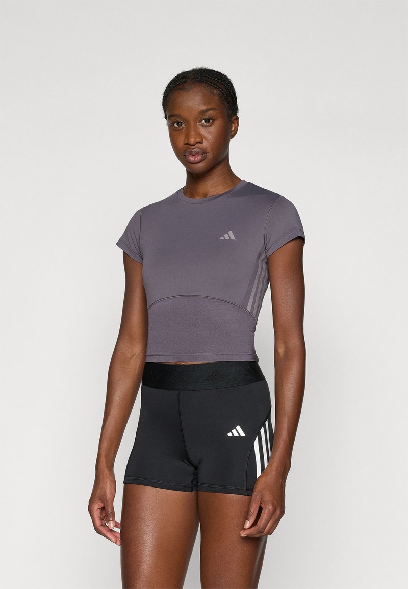 Lühikeste varrukate ja ümmarguse kaelusega lilla sportlik crop top halli logoga, koos mustade lühikeste pükste ja valgete triipudega, millel on veniv vöökoht.