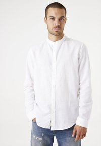 Camisa de lino blanca de manga larga con cuello de banda, cierre delantero de botones y dobladillo ligeramente curvado. Combinada con unos vaqueros desgastados de color azul.
