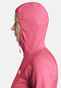 Veste à capuche zippée rose avec un tissu texturé. Elle présente un col haut, une capuche ajustée et un logo sur la poitrine. Vue de profil.
